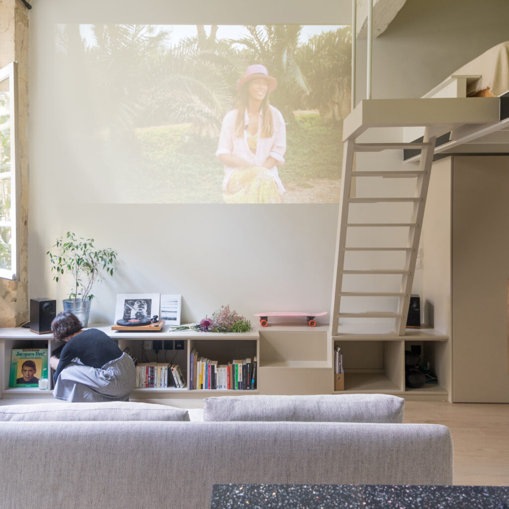 image d'un petit salon avec une mezzanine acccesible par un meuble bas par une echelle il y a un grand mur blanc qui sert de surface pour un vidéo projecteur