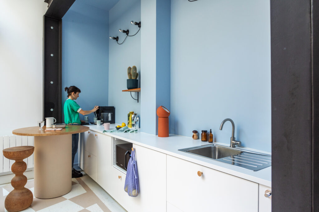 une cuisine blanche avec un mur bleu ciel une femme preprare un café. le sol est en damier beige et blanc peint en trompe l'oeil